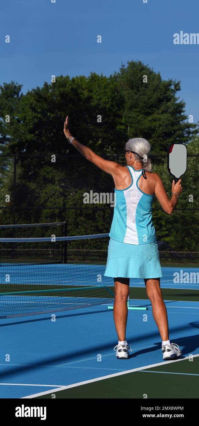 Senior female stands on the serving line with her hand up, instucting others to wait.  She is wearing a turquoise tennis outfit. Stock Photo