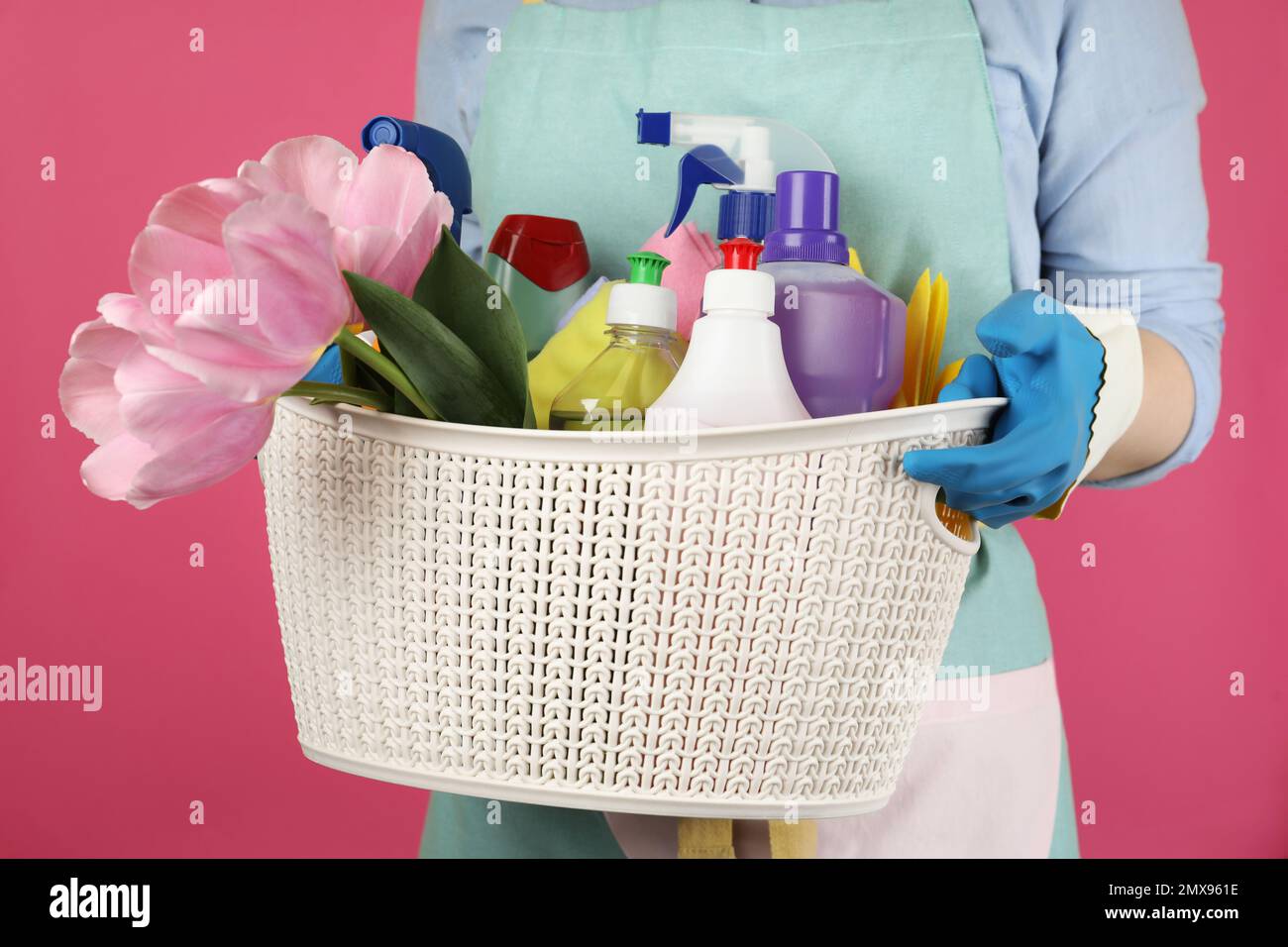 Woman holding basket with spring flowers and cleaning supplies on pink background, closeup Stock Photo