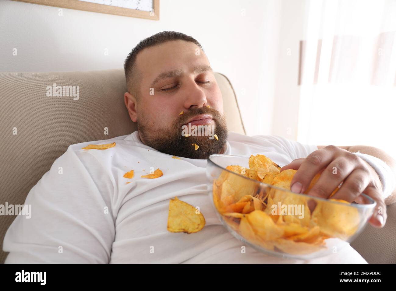 Lazy overweight man with chips sleeping on sofa at home, closeup Stock Photo
