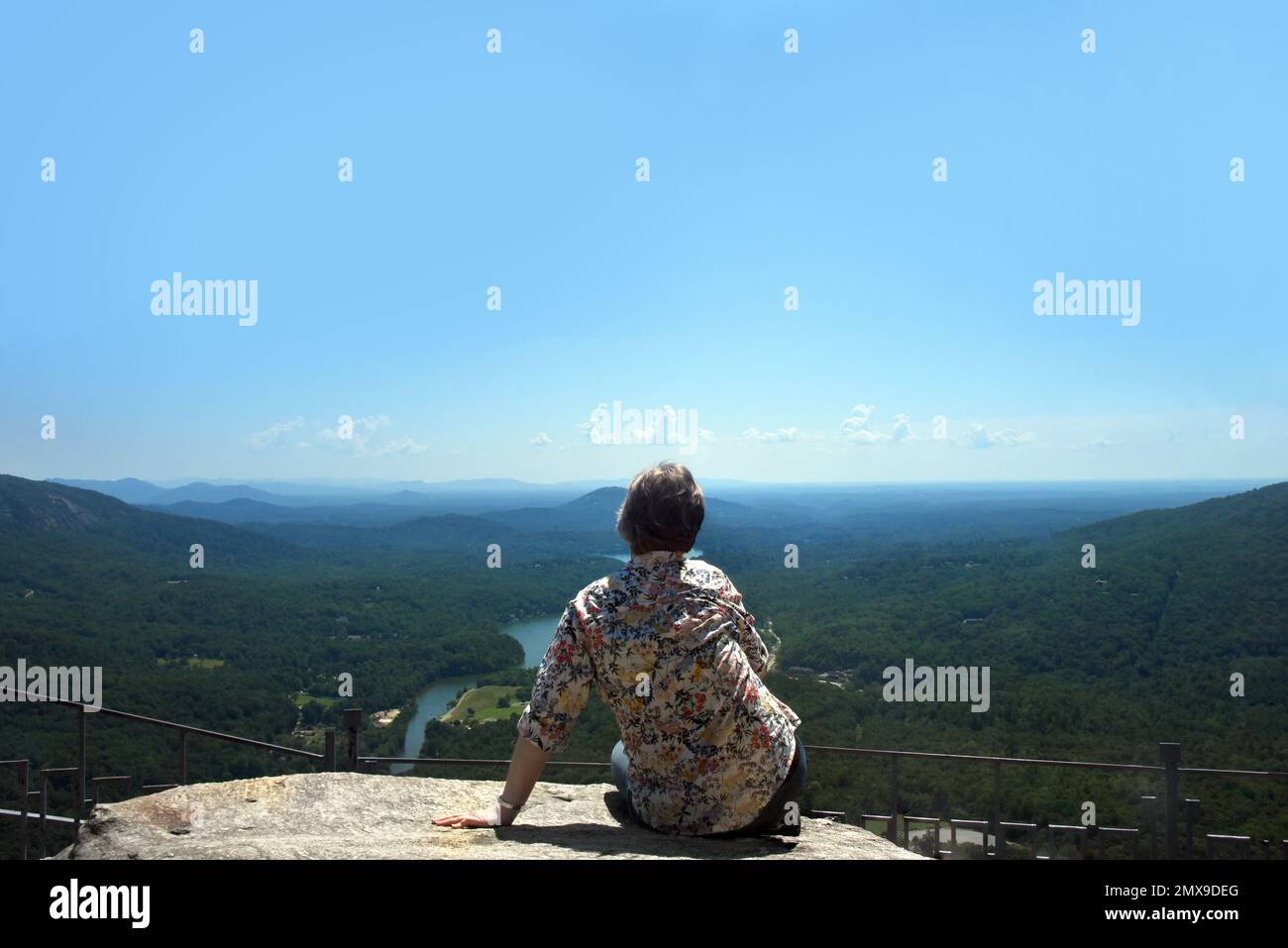 Visitor sits absorbing the view at the top of Chimney Rock, Chimney Rock State Park, North Carolina.  Visitor is a senior citizen and is wearing jeans Stock Photo