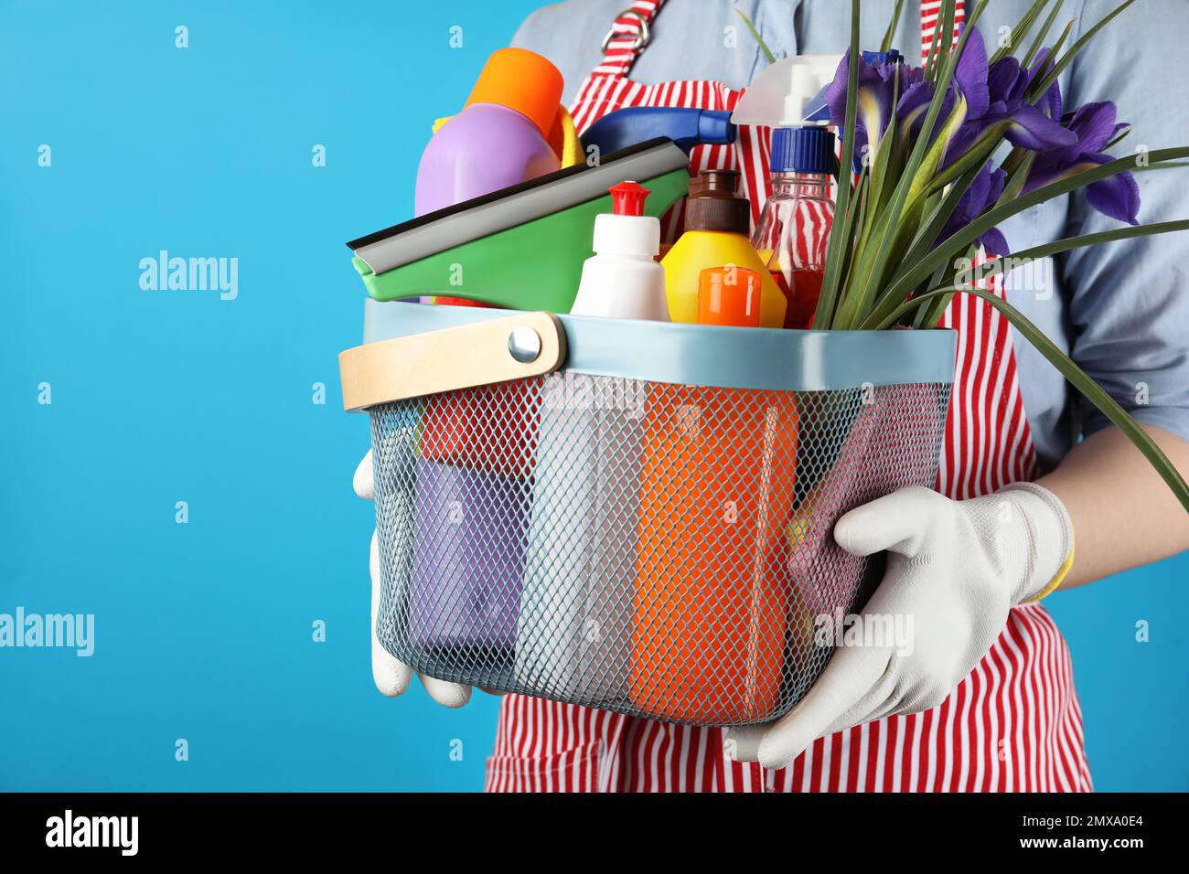 Woman holding basket with spring flowers and cleaning supplies on light blue background, closeup Stock Photo