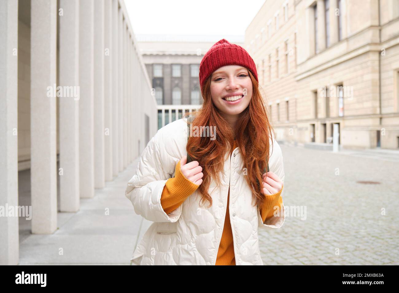 Female tourist in red hat with backpack, sightseeing, explores historical landmarks on her trip around europe, smiling and posing on street. Stock Photo