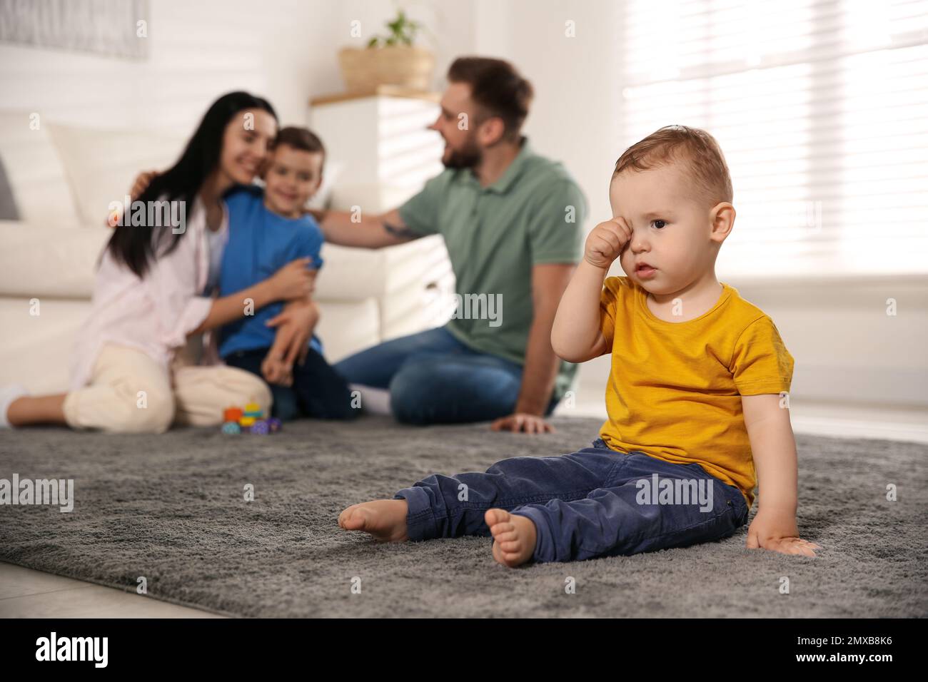 Unhappy baby sitting alone on floor while parents spending time with his elder brother at home. Jealousy in family Stock Photo