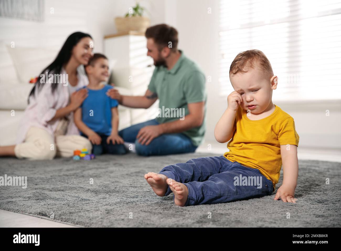 Unhappy baby sitting alone on floor while parents spending time with his elder brother at home. Jealousy in family Stock Photo
