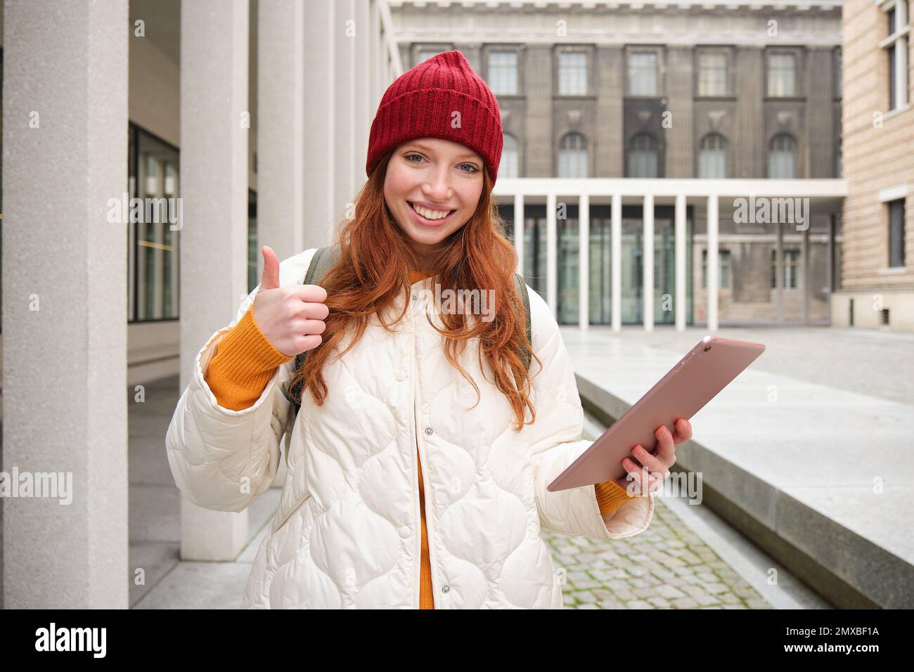 Stylish ginger girl, tourist walks with digital tablet around city, woman connects to iternet on her gadget, looking up information, texting message. Stock Photo