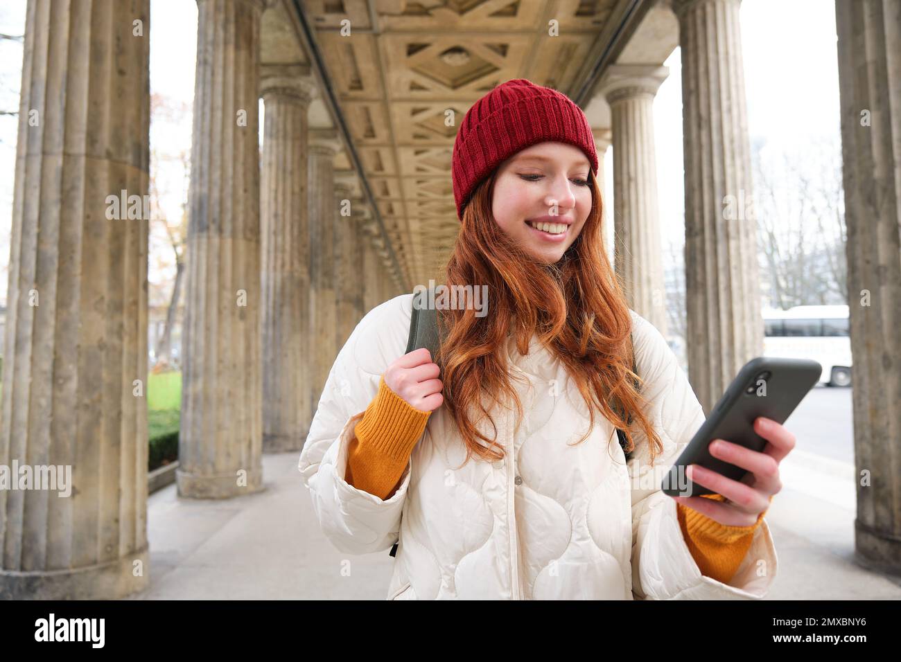 Mobile broadband and people. Smiling redhead 20s girl with backpack, uses smartphone on street, holds mobile phone and looks at application. Stock Photo