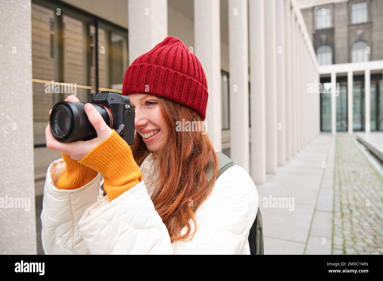 Smiling redhead girl photographer, taking pictures in city, makes photos outdoors on professional camera. Young talent and hobby concept Stock Photo