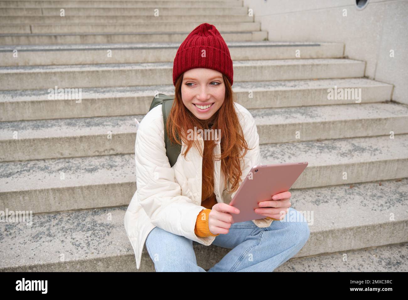 Young stylish girl, redhead female students sits on stairs outdoors with digital tablet, reads, uses social media app on gadget, plays games while wai Stock Photo
