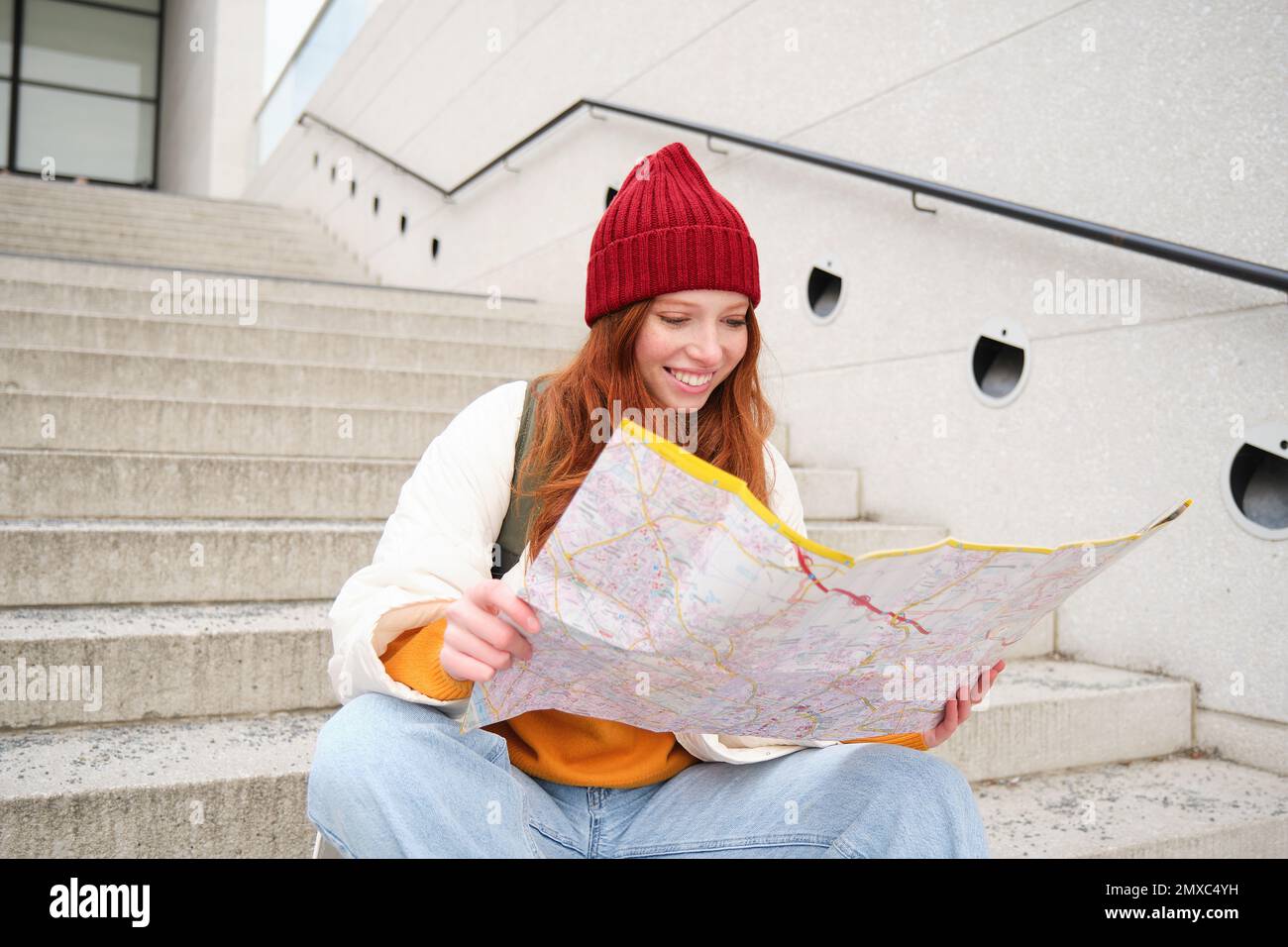 Young smiling redhead girl, tourist sits on stairs outdoors with city paper map, looking for direction, traveller backpacker explores city and looks f Stock Photo