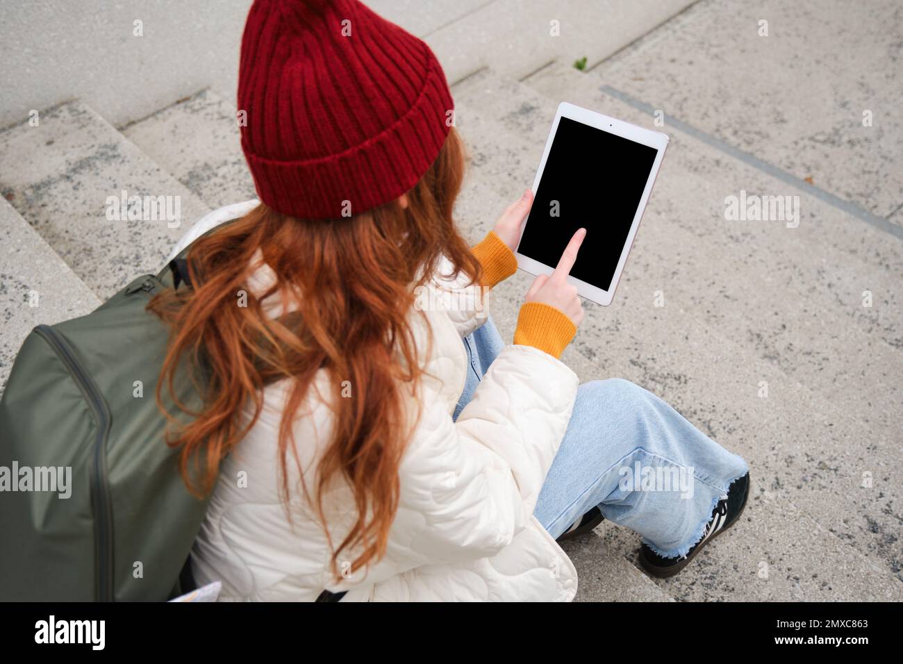 Rear view of redhead girl touches digital tablet screen, touchpad, texts message, uses internet application on gadget, sits on stairs outdoors. Stock Photo