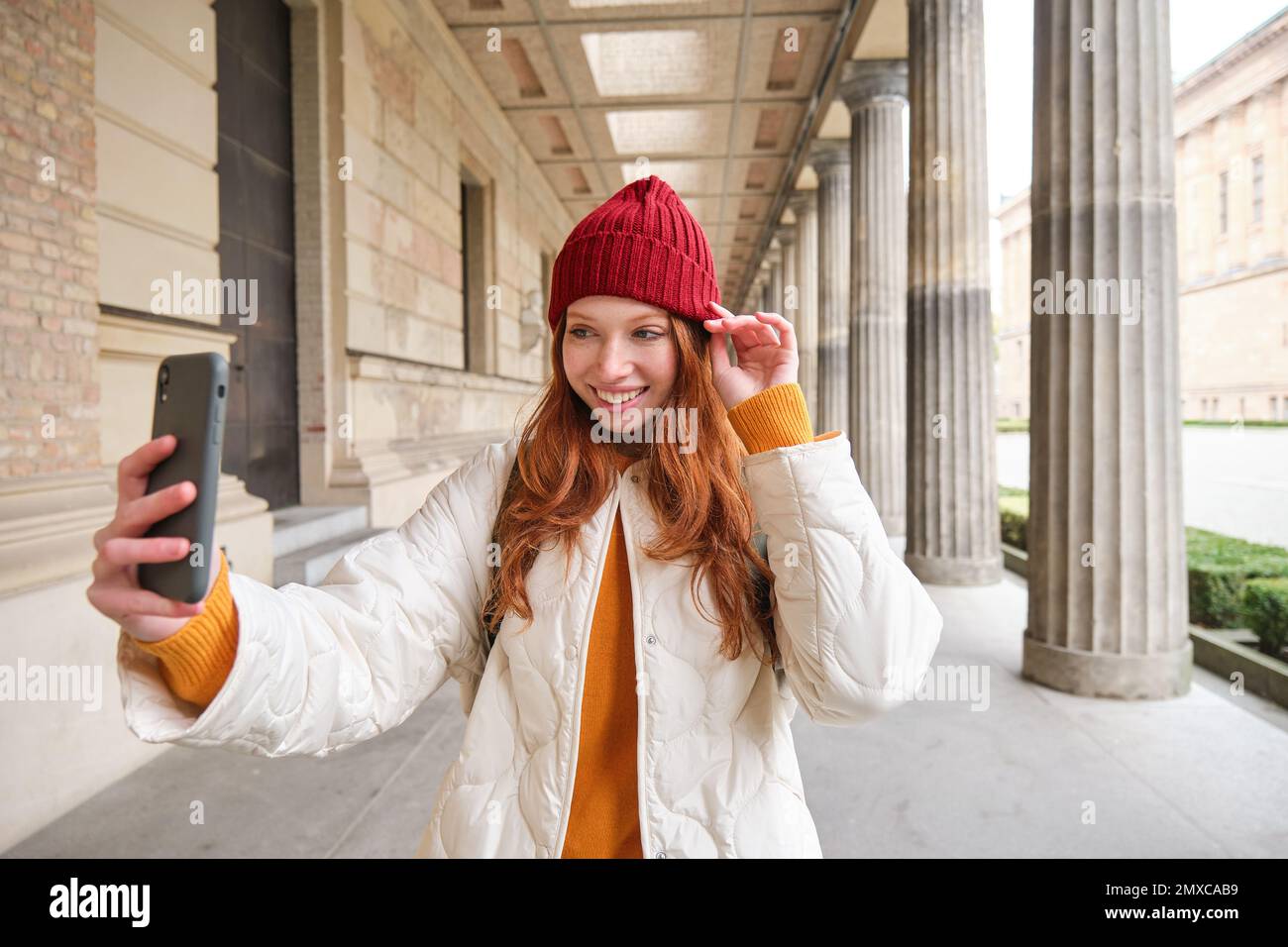 Stylish redhead girl tourist, takes selfie in front of tourism attraction, makes photo with smartphone, looks at mobile camera and poses. Stock Photo