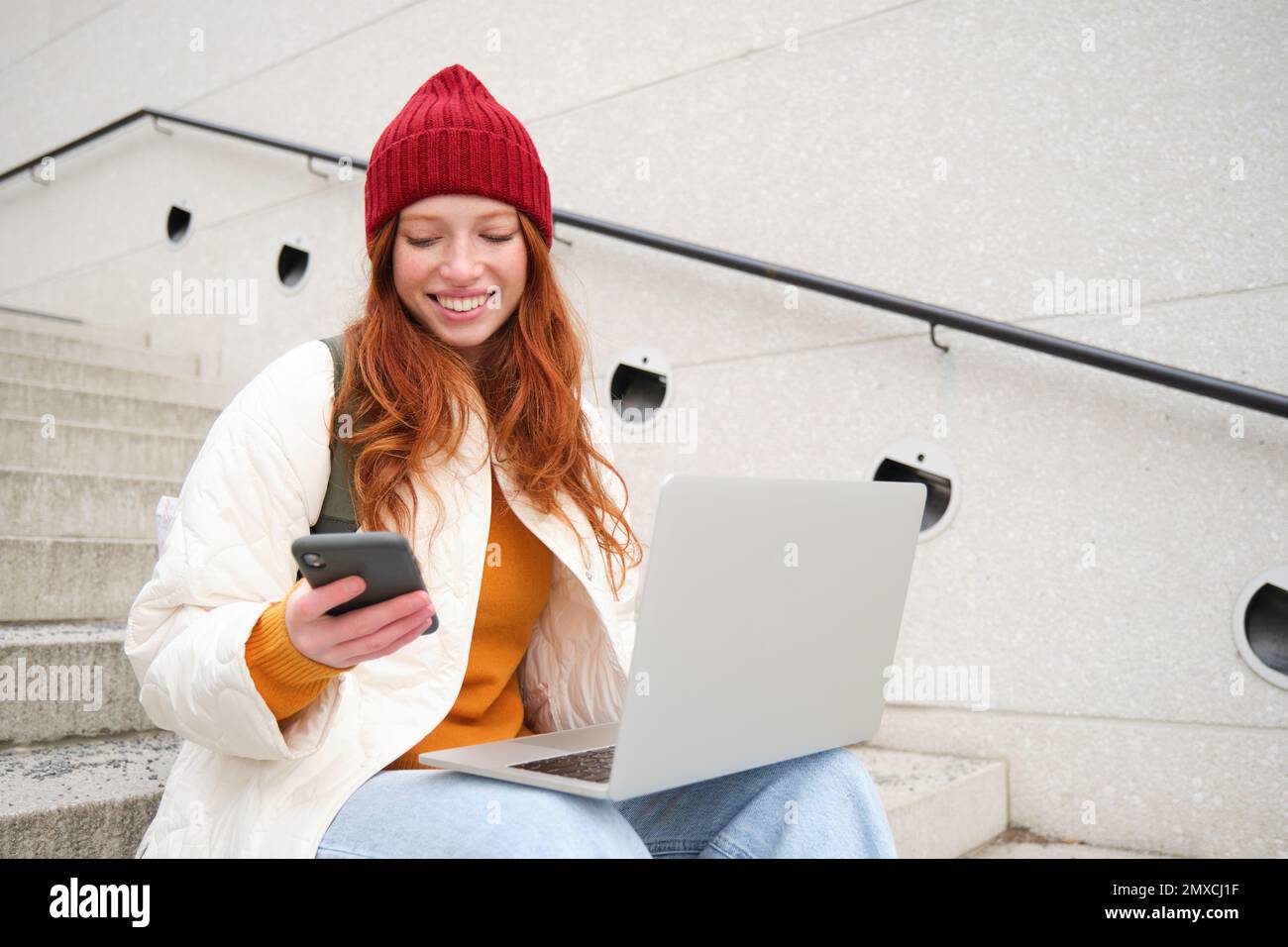 Young tourist, girl booking hotel room on laptop, enters confirmation code on smartphone, sitting with computer and phone on stairs outdoors. Stock Photo