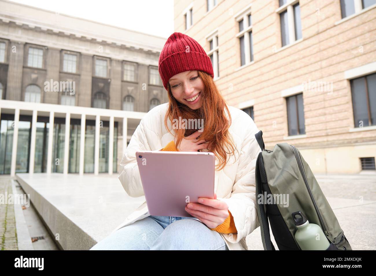 Redhead girl smiles, sits outdoors near building with digital tablet, thermos and backpack, connects to public internet and searches smth online on he Stock Photo