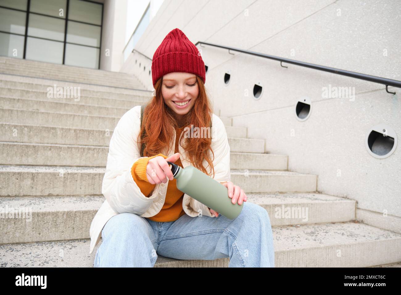 Happy redhead woman, tourist with thermos, drinks her hot tea, coffee from travel flask, restests during her travelling in city and sightseeing Stock Photo