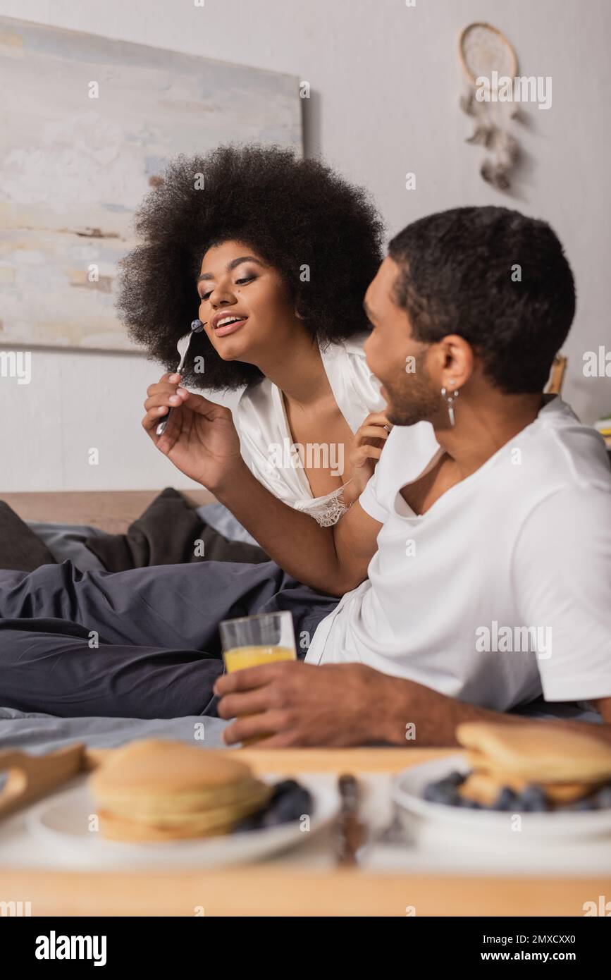 young african american man holding orange juice and feeding sexy girlfriend with berry during breakfast Stock Photo