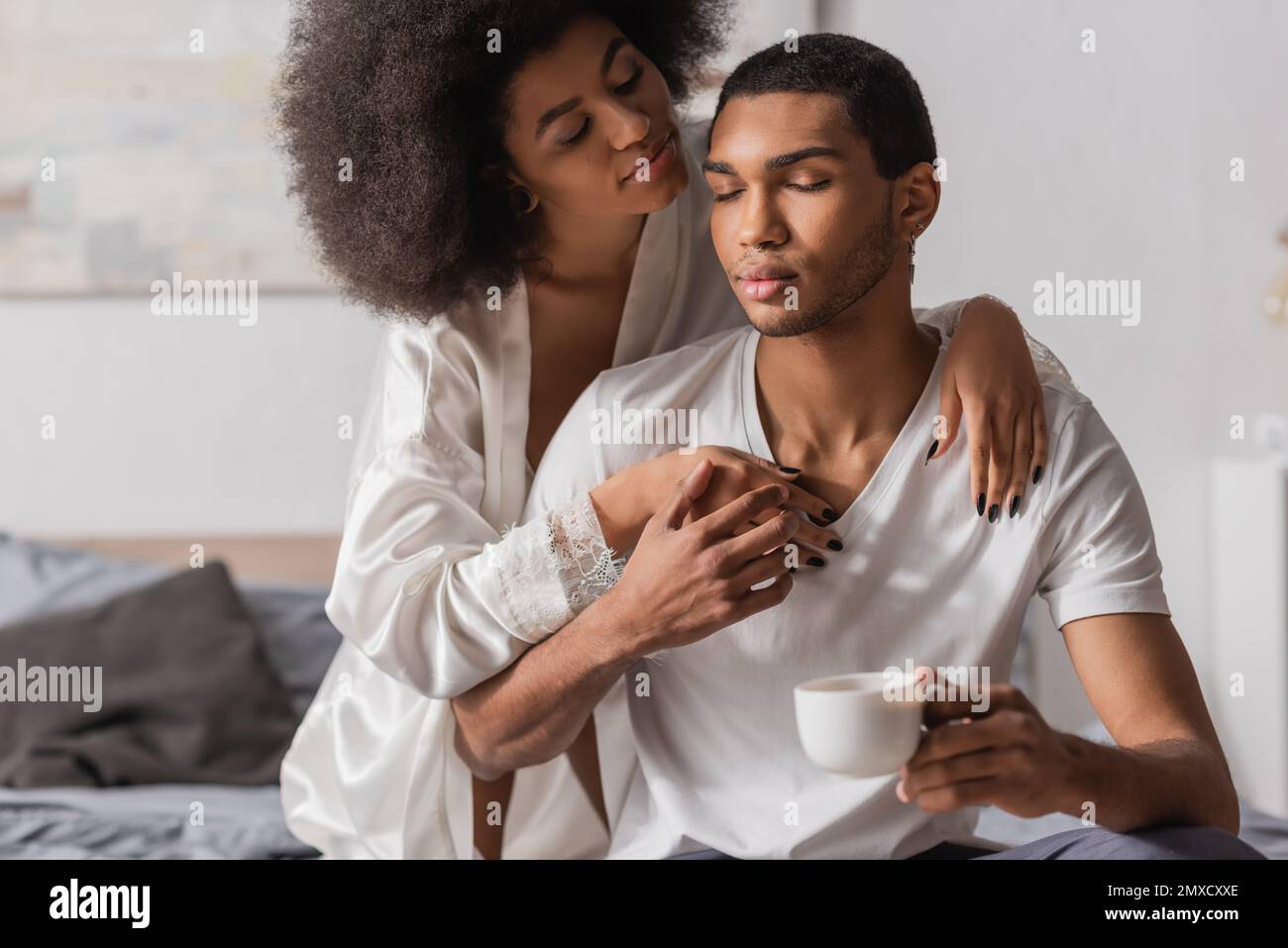 sensual african american woman in white satin robe embracing young man holding coffee cup with closed eyes Stock Photo