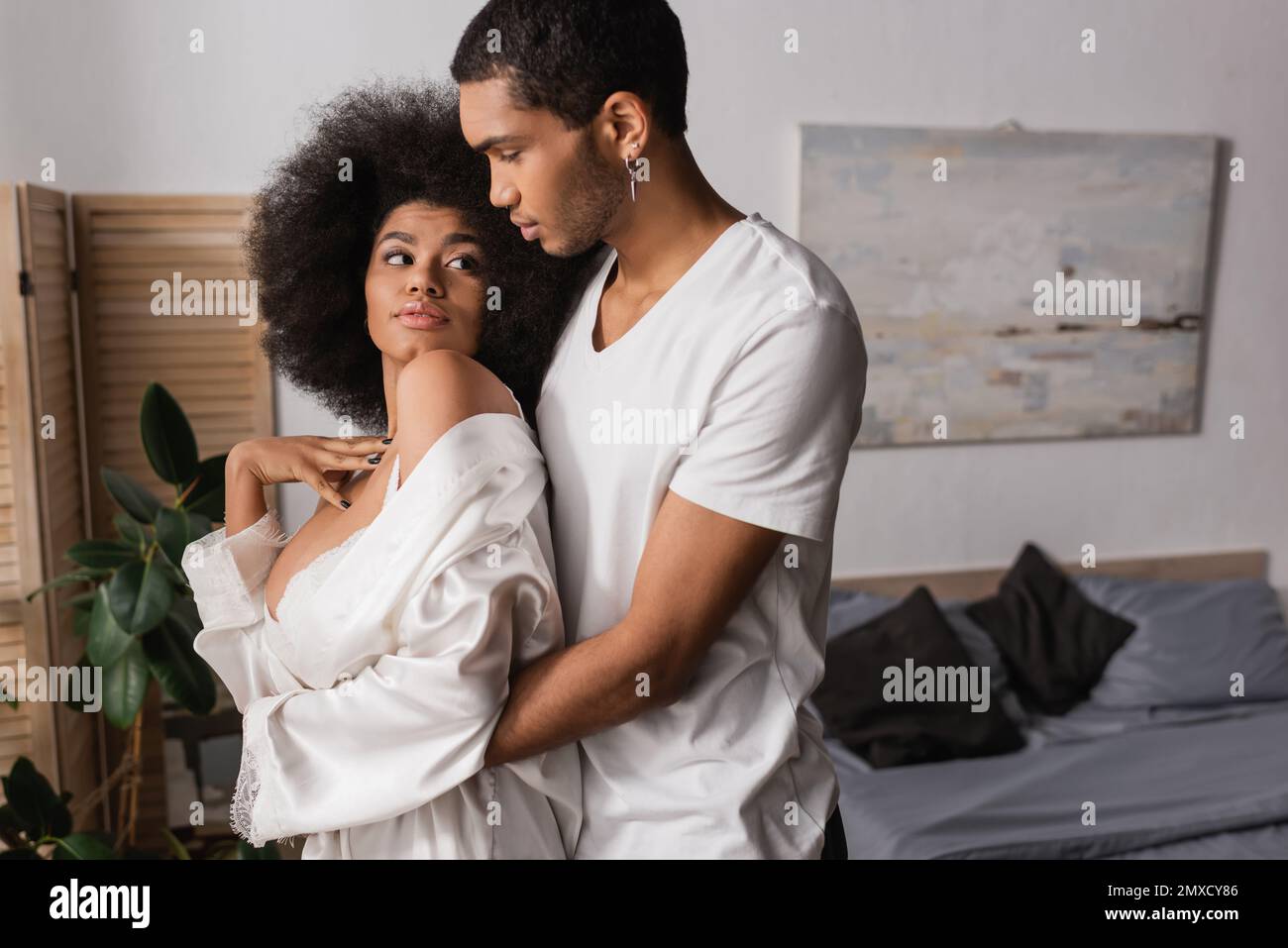 young african american man hugging sexy woman in white silk robe in bedroom Stock Photo