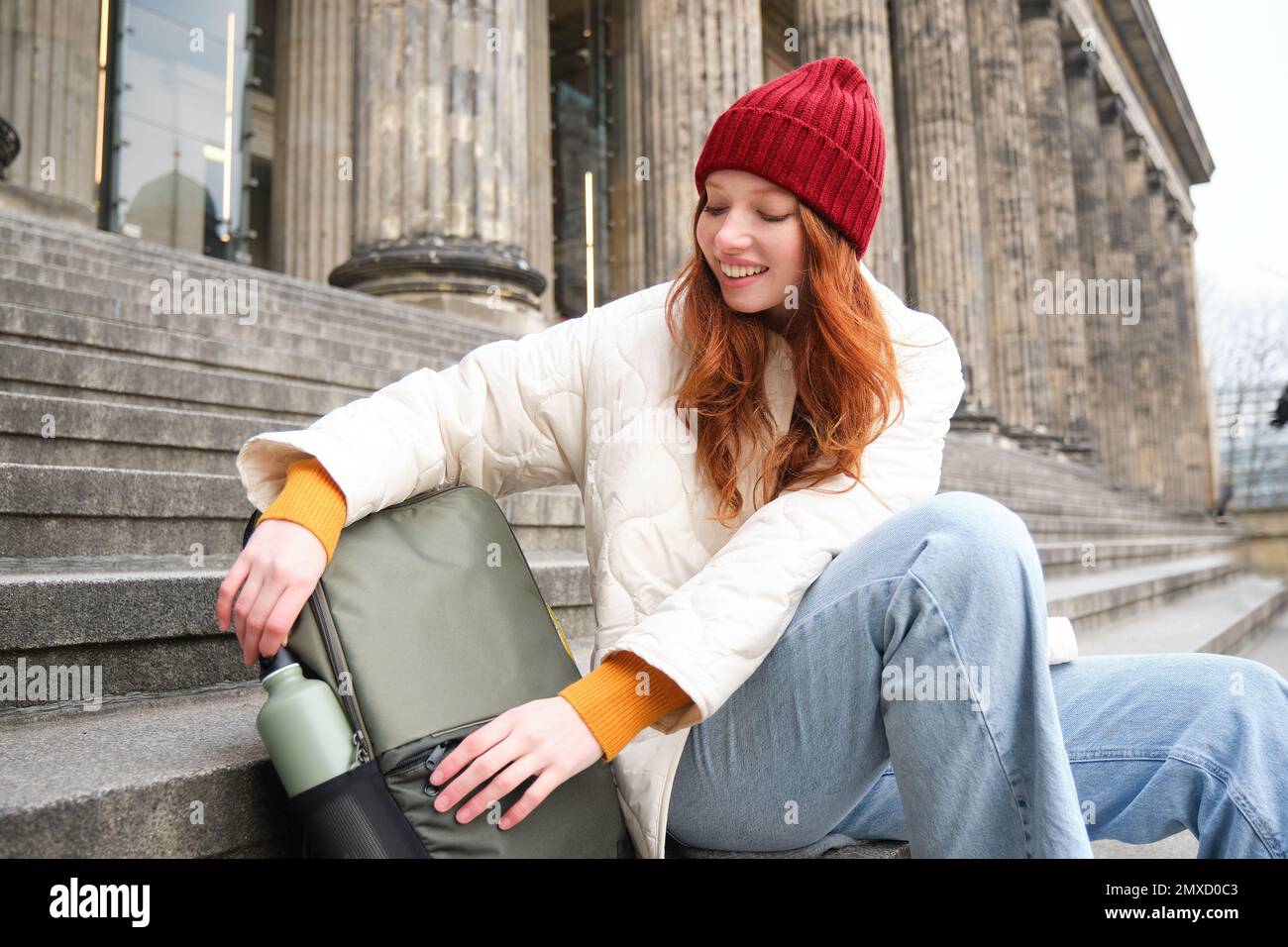 Smiling tourist, girl sits on stairs, rests on staircase, takes thermos from backpack, drinks hot coffee from flask Stock Photo