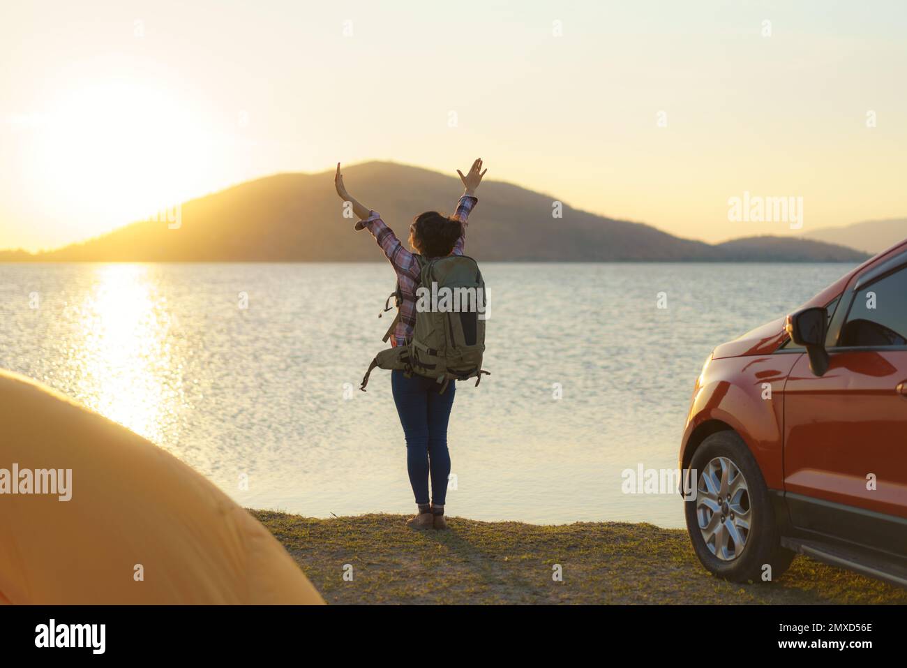 Asian backpacker woman raising her hands happy to arrive at her destination where she is camping with a lake in the background during sunset. Stock Photo