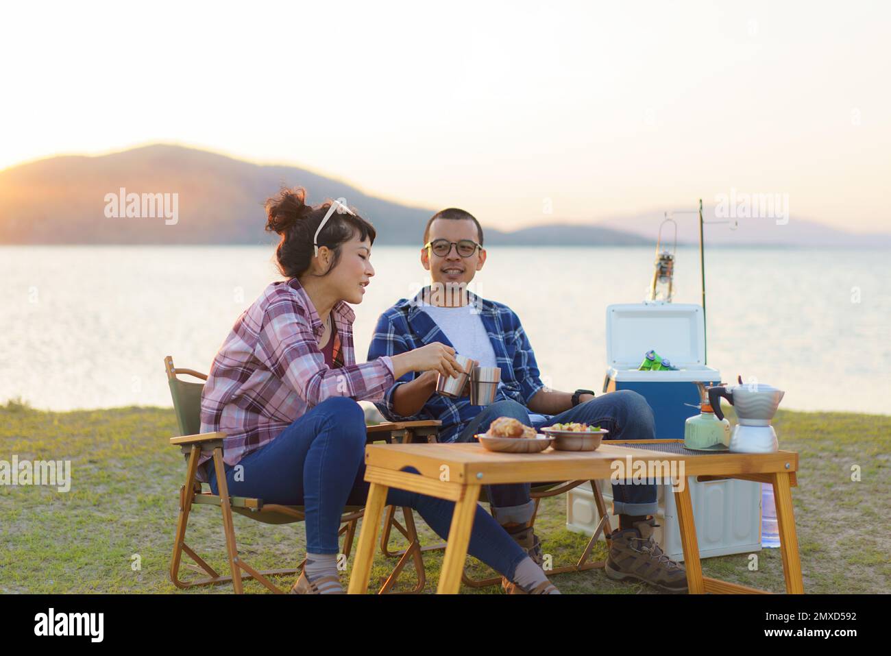 Asian couple having a cup of coffee or water at their camping site with lake in the background during sunset Stock Photo