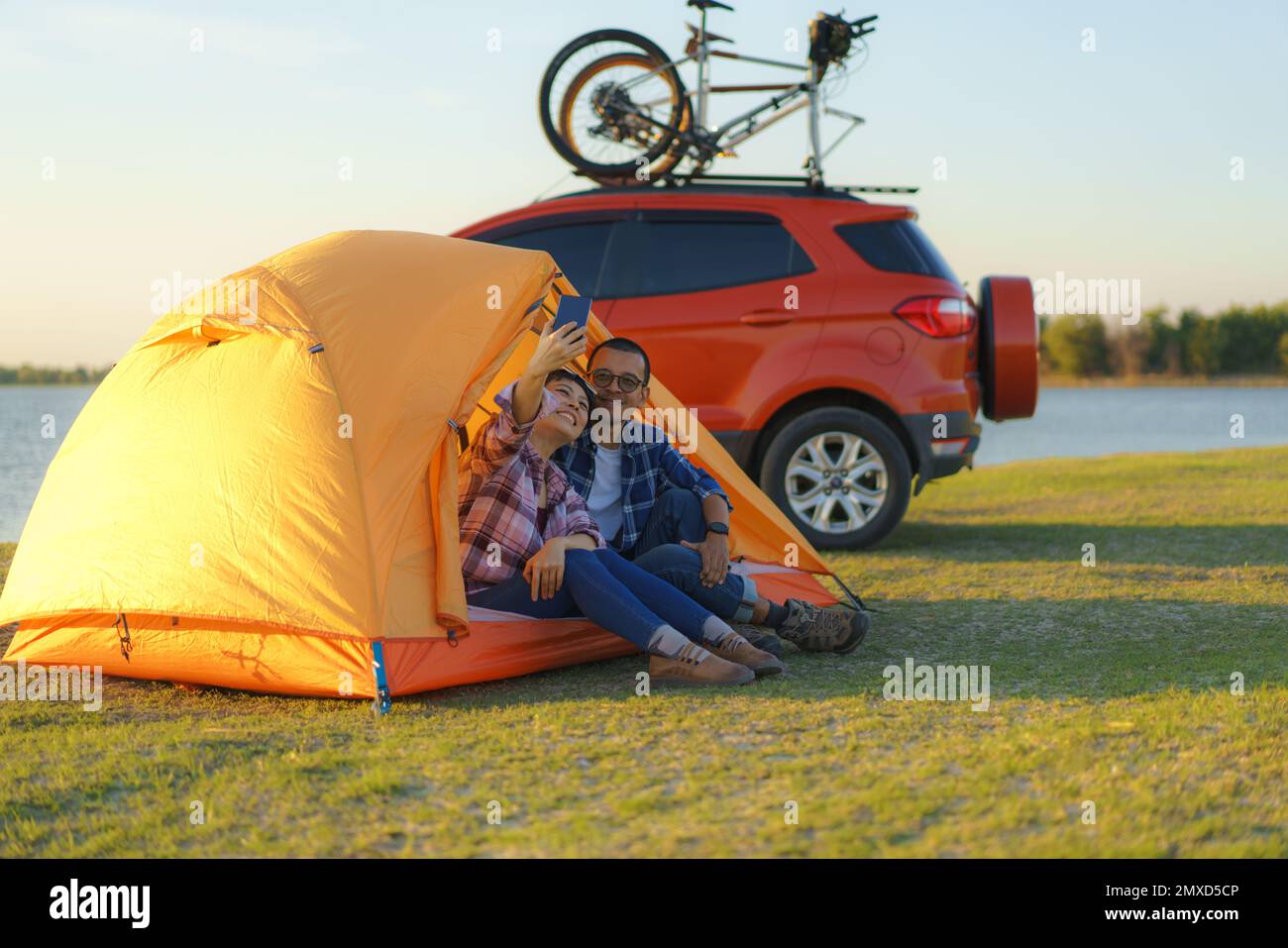 Young Asian couple taking selfie by smartphone camera by sitting in a tent while camping with the lake in the background during sunset Stock Photo