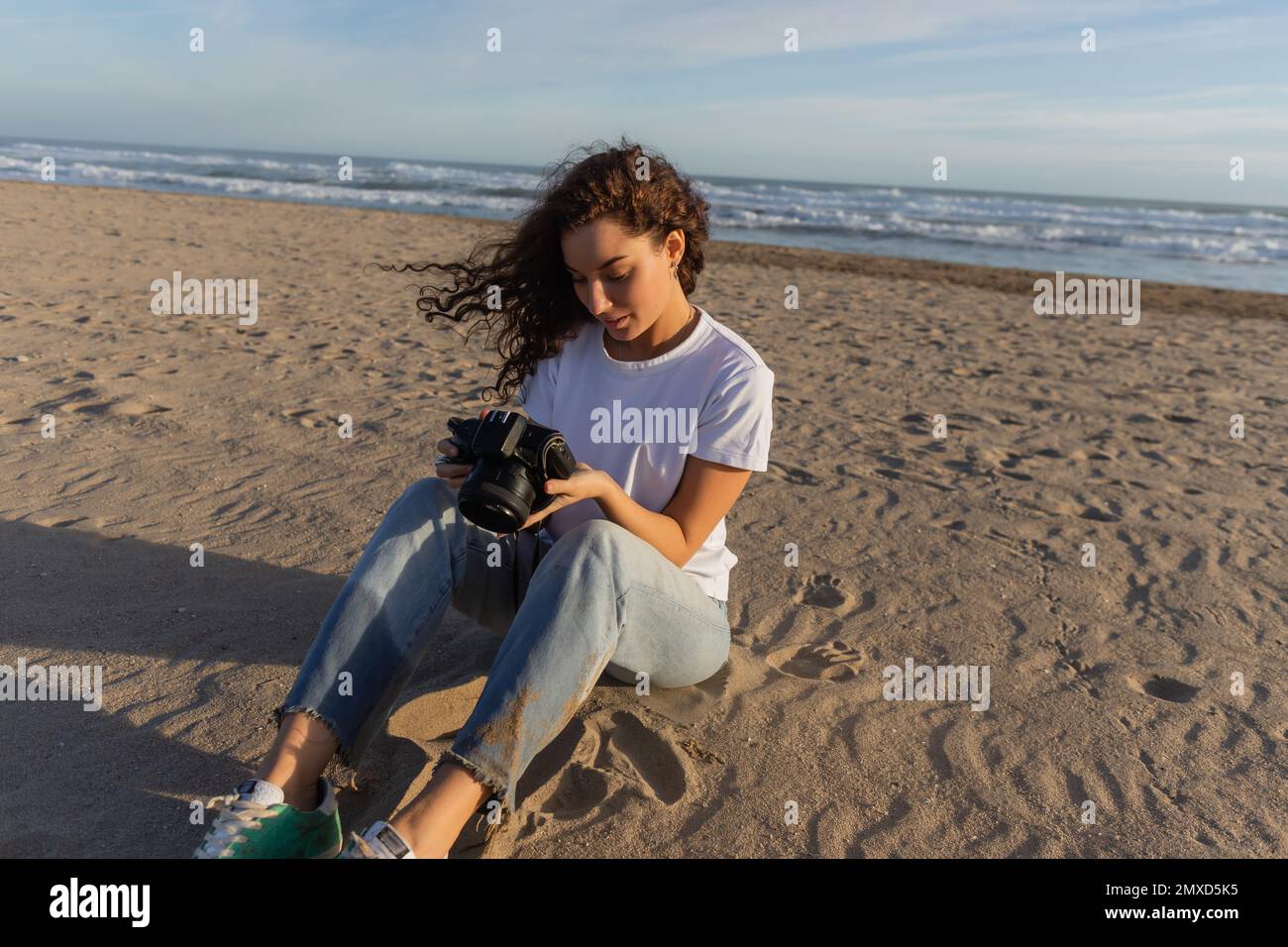 curly woman sitting in blue jeans and white t-shirt sitting with digital camera on beach in Spain Stock Photo