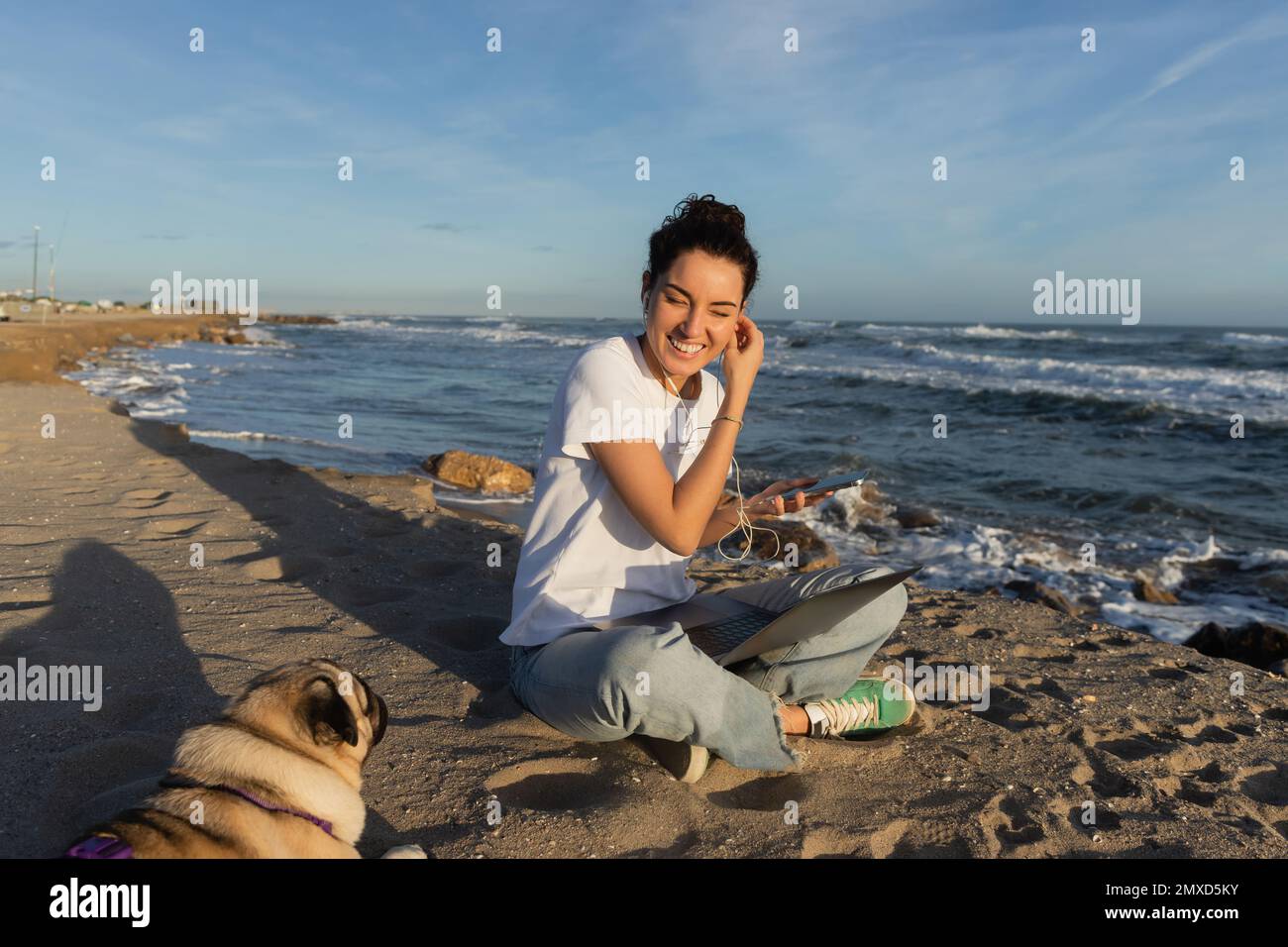 happy woman wearing wired earphones while holding smartphone near laptop and pug dog on beach in Barcelona Stock Photo