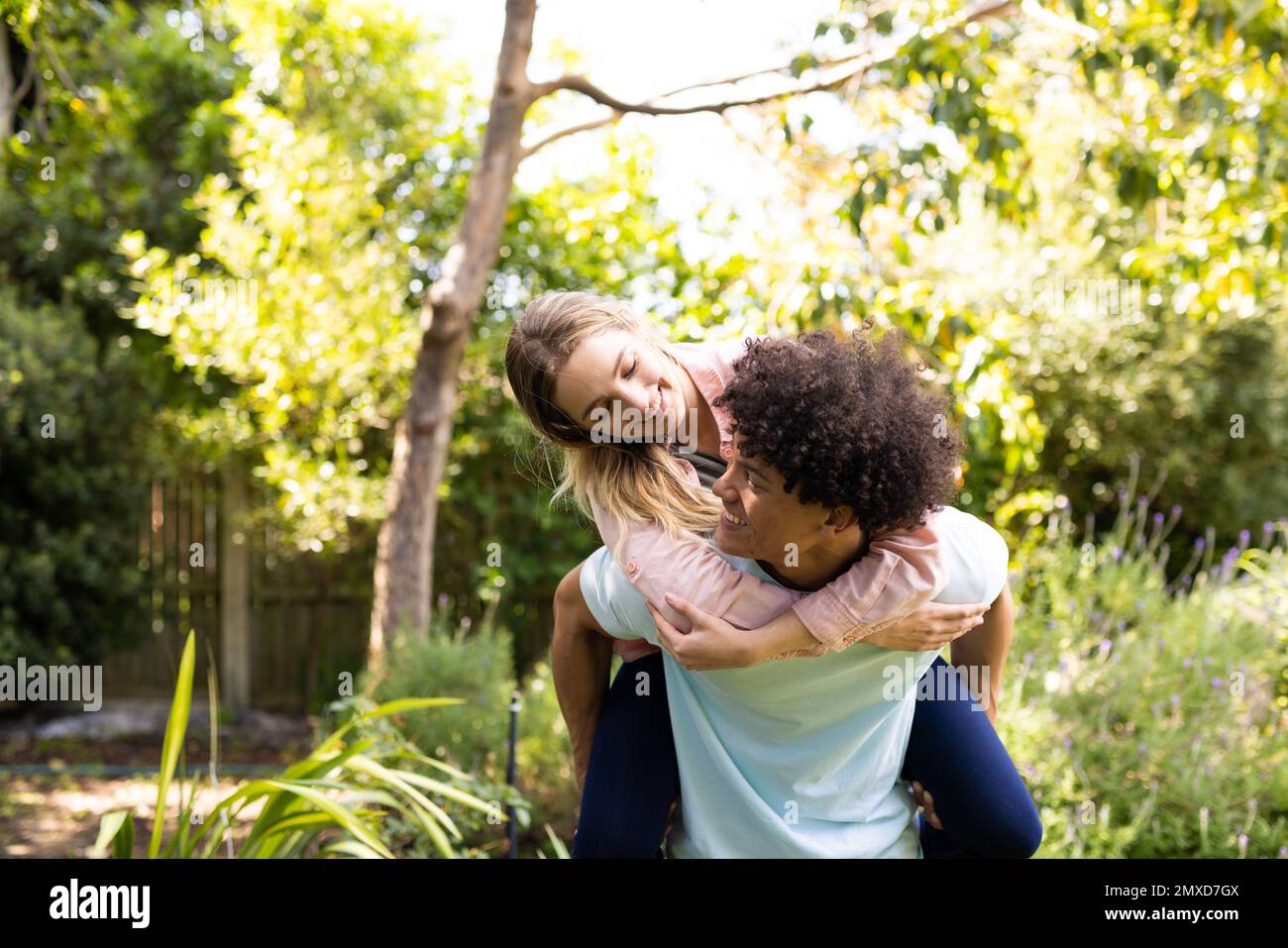 Happy diverse couple having fun piggybacking in sunny garden, copy space Stock Photo