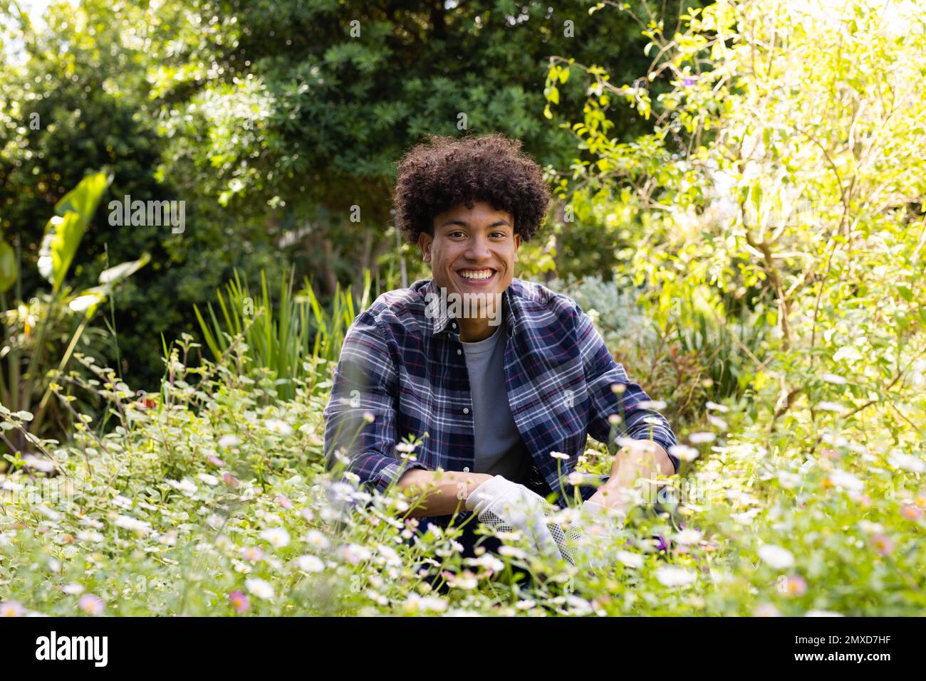 Portrait of happy biracial man gardening, sitting amongst flowers in sunny garden Stock Photo