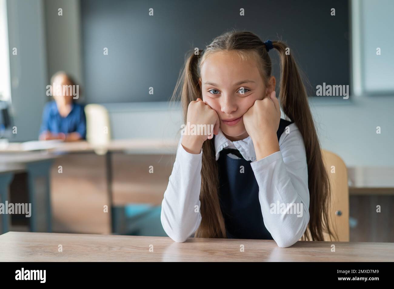 Little caucasian girl is bored at the lesson at school. The schoolgirl is sitting at her desk and the teacher is sitting in the background. Stock Photo