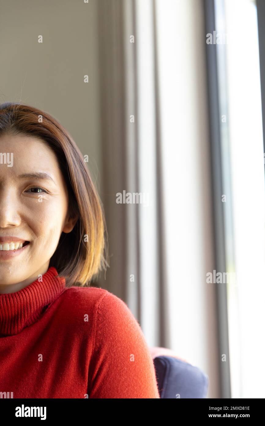 Vertical half face portrait of happy asian woman sitting by window, smiling to camera, copy space Stock Photo