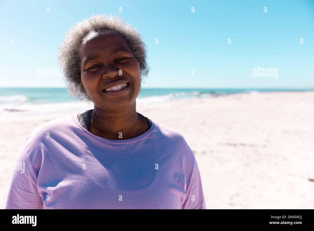 Smiling african american senior woman with short gray hair standing on beach against sea and sky Stock Photo