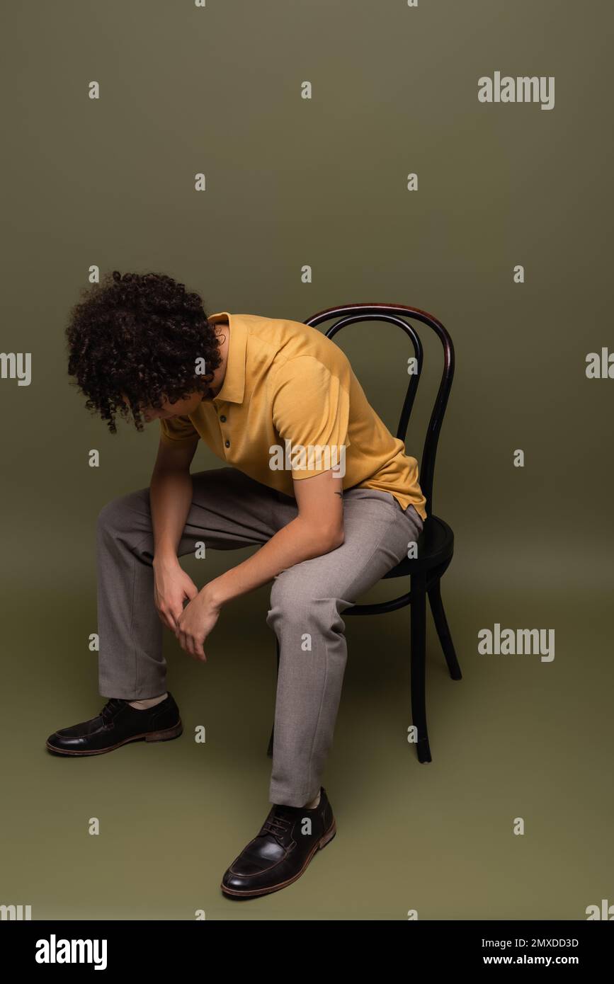 full length of african american man in yellow polo shirt and black leather shoes sitting on chair with bowed head on grey green background,stock image Stock Photo