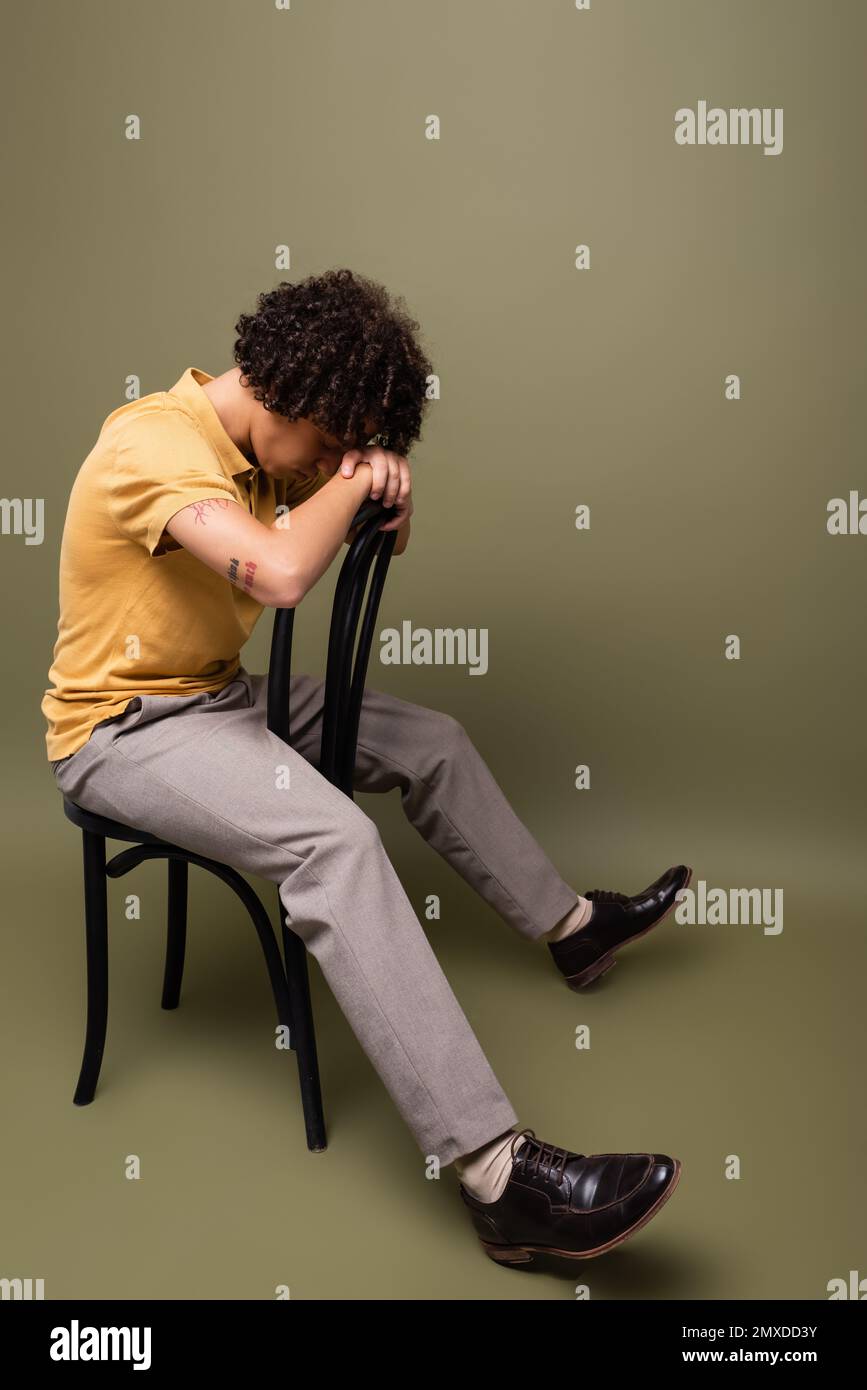 full length of tattooed african american man in yellow polo shirt and pants sitting on chair with bowed head on grey green background,stock image Stock Photo