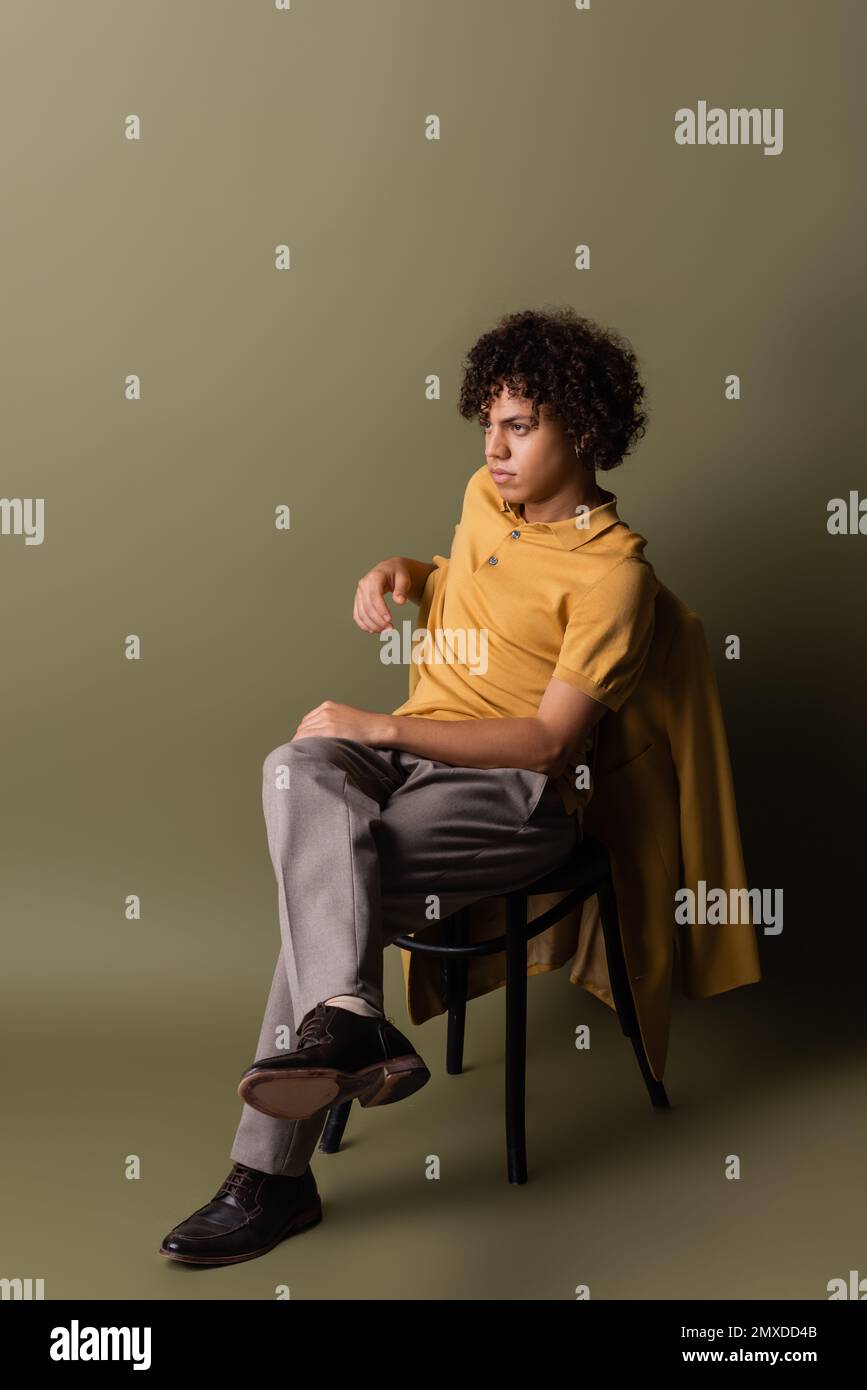full length of african american man in yellow polo shirt sitting with crossed legs and looking away on grey green background,stock image Stock Photo