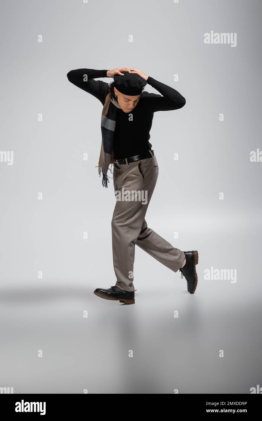 full length of african american man in black beret and sweater with plaid scarf walking with hands above head on grey background,stock image Stock Photo
