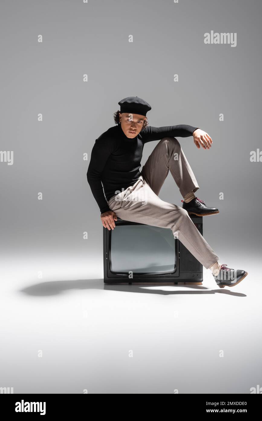 full length of african american man in stylish beret and black pullover sitting on retro tv and looking at camera on grey background,stock image Stock Photo