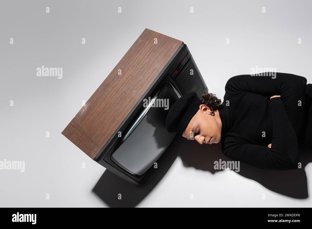 top view of african american man in black pullover and beret lying with crossed arms near retro tv set on grey background,stock image Stock Photo