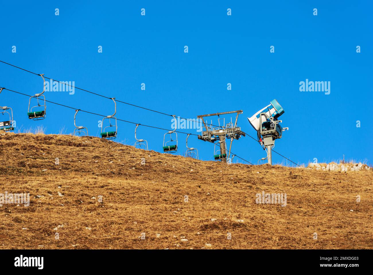 Snow cannon or snowmaking system and an empty chairlift in winter on a brown meadow, ski slope, without snow due to the too hot. Veneto, Italy. Stock Photo