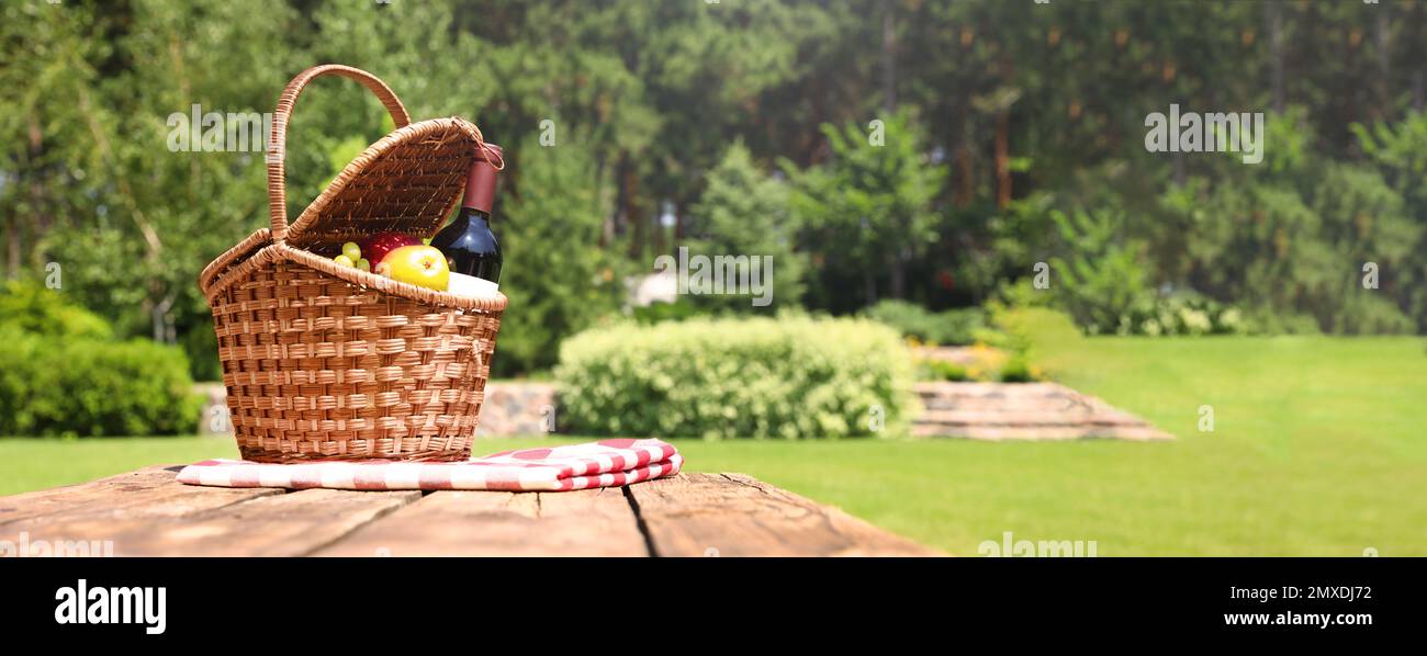 Picnic basket with fruits, bottle of wine and checkered blanket on wooden table in garden, space for text. Banner design Stock Photo