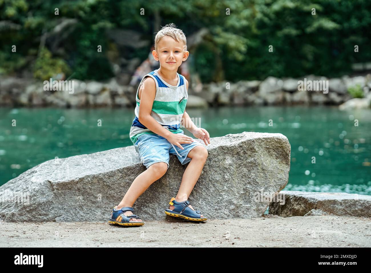 Excited child sits on rock near turquoise pond illuminated by sunlight. Happy preschooler boy smiles and enjoys exploring magic places in resort town Stock Photo