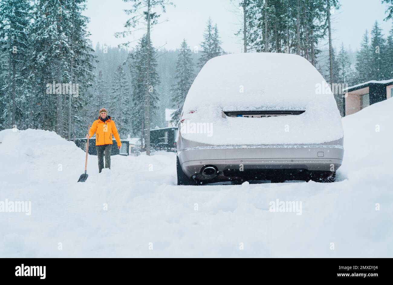 Car covered with snow as a huge snowdrift on the countryside forest home driveway. Smiling man with a shovel going to remove snow from the path and cl Stock Photo