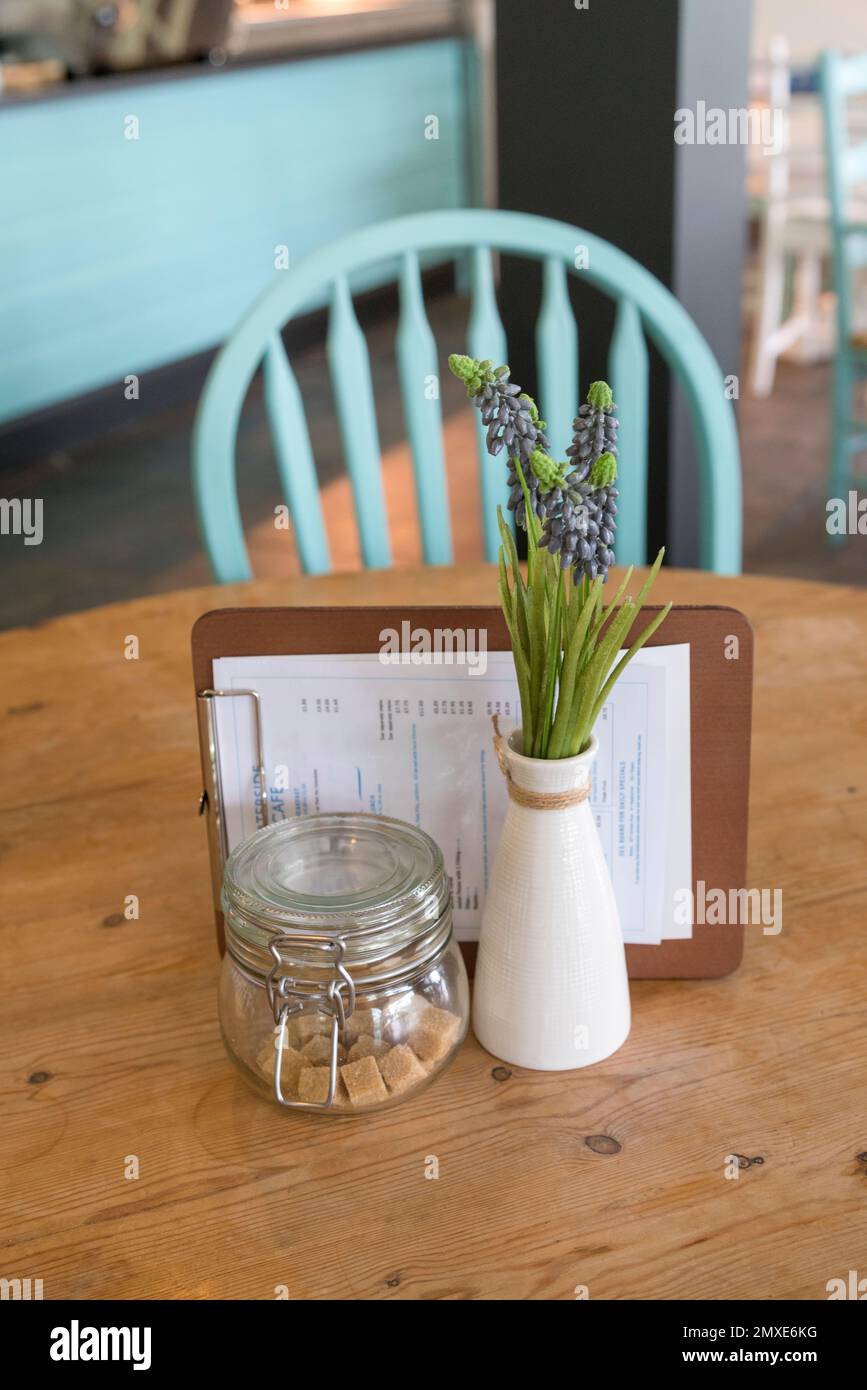 Close up of a clean, empty cafe table with a vase of lavender - an empty turquoise wooden ladder back chair is shown in the defocussed background. Stock Photo