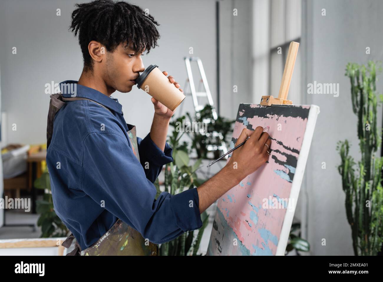 Young african american artist drinking coffee to go and painting on canvas in workshop,stock image Stock Photo