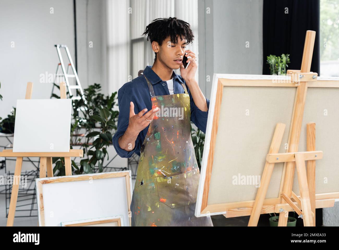 African american artist in dirty apron talking on smartphone near canvas in studio,stock image Stock Photo