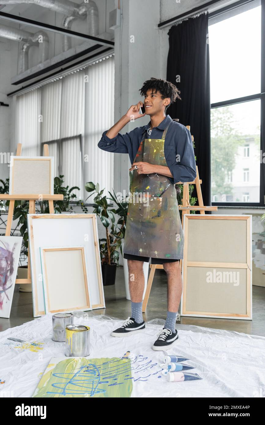 Smiling african american artist in apron talking on smartphone near paints in studio,stock image Stock Photo