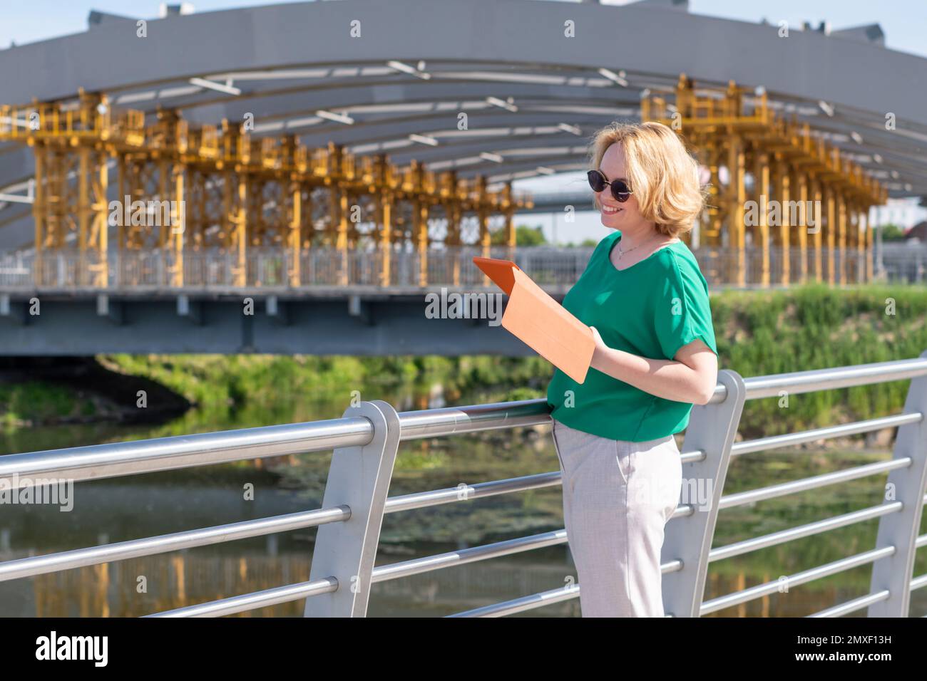 A smiling woman in sunglasses holding a tablet in her hands, reading news, using applications on the street.  An attractive girlreading a book on stre Stock Photo