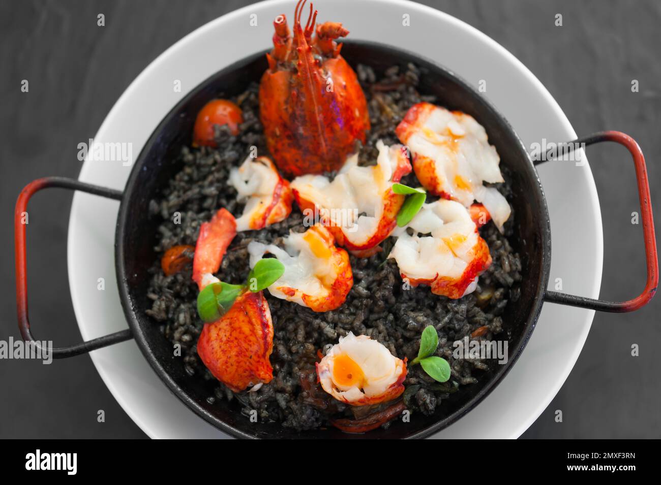 Solo paella with lobster slices, tomato chunks, squid ink sauce, herbs, saffron. Hot dish on a white plate. Meal on a black table. Modern food plating. Stock Photo