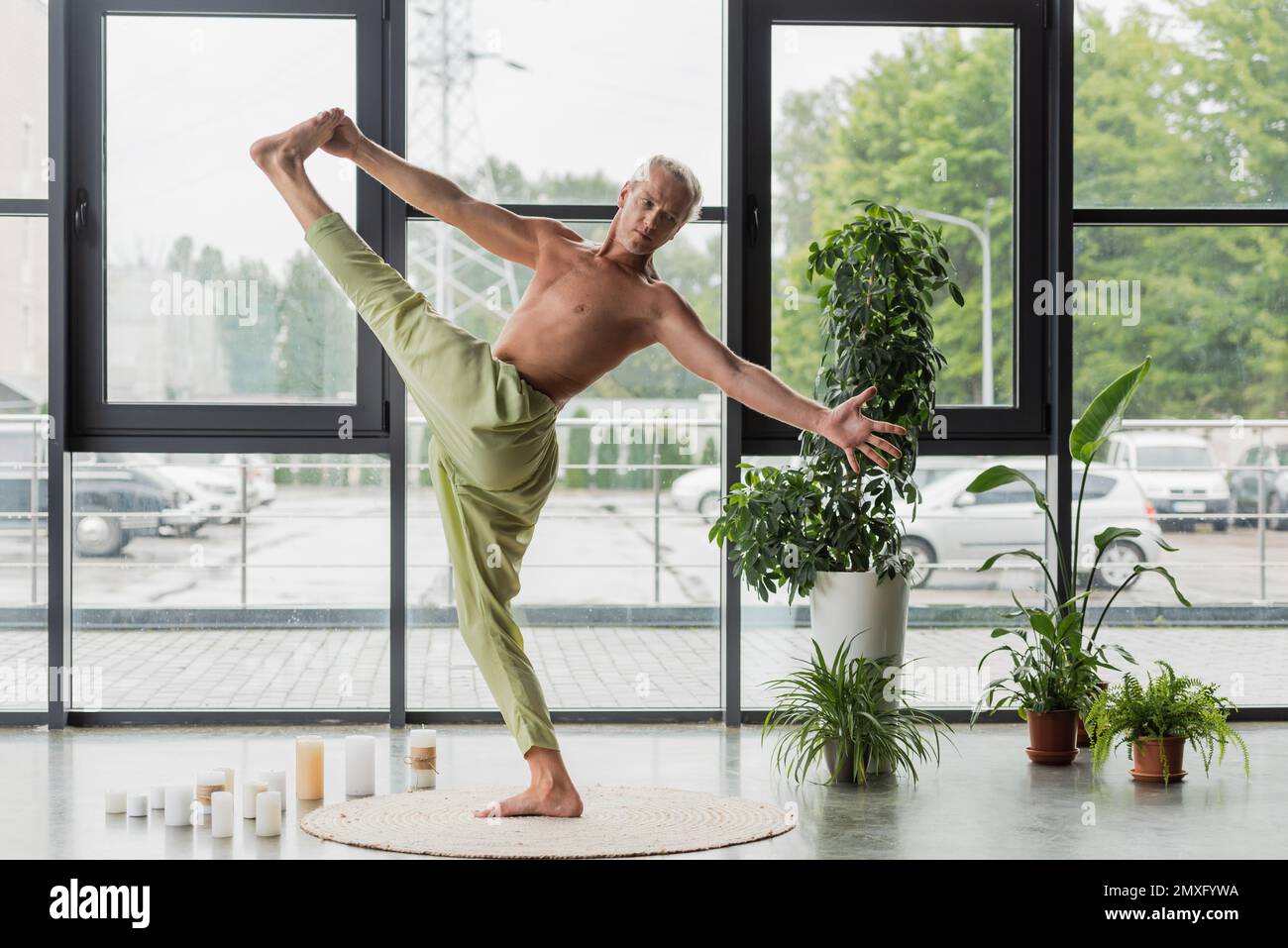 shirtless man in green pants doing hand to big toe pose near candles and plants in yoga studio,stock image Stock Photo