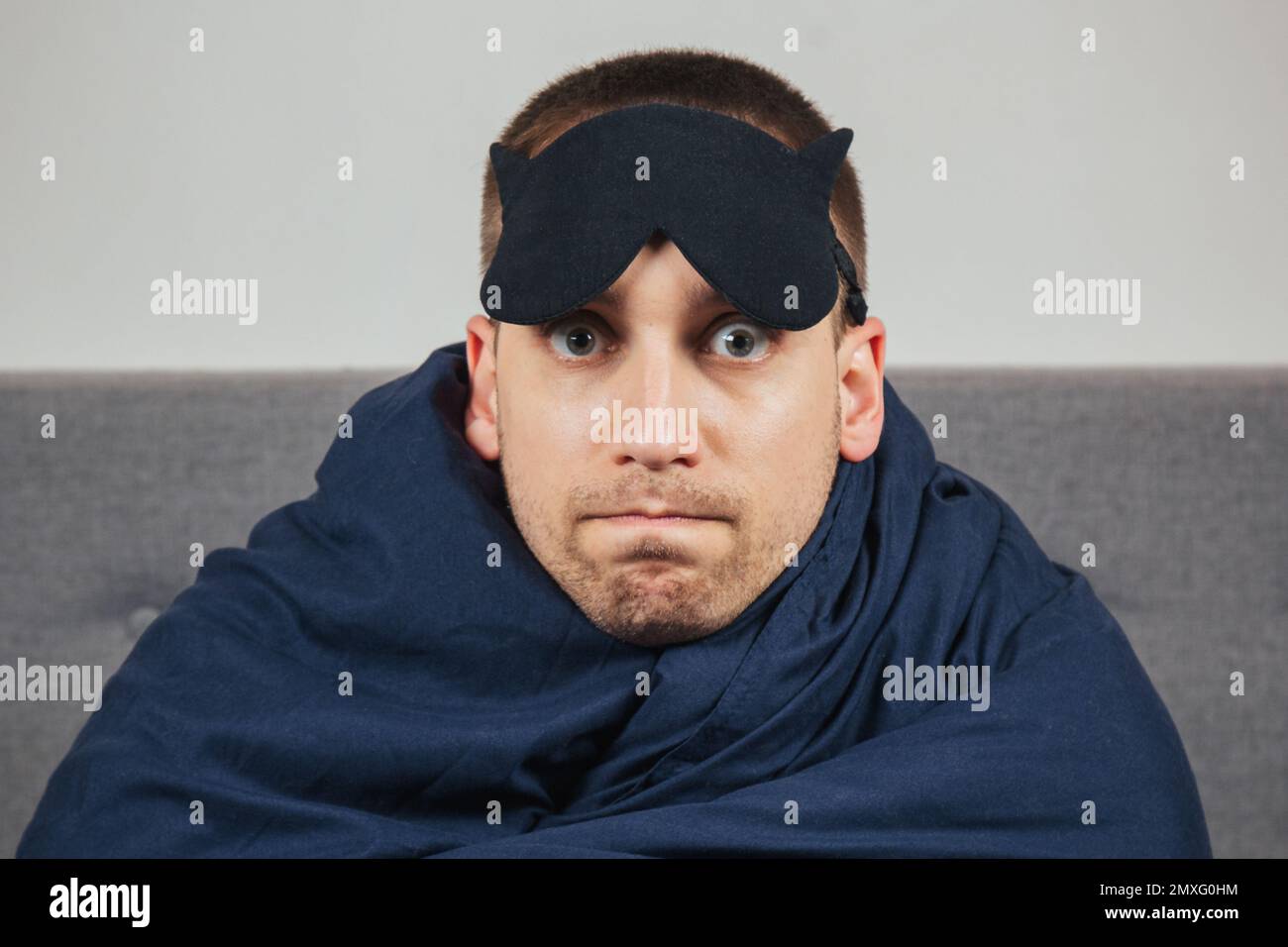 Embarrassed young man in a blanket at home wears a sleep mask while relaxing at home, studio portrait. Relax, lifestyle concept in a good mood Stock Photo